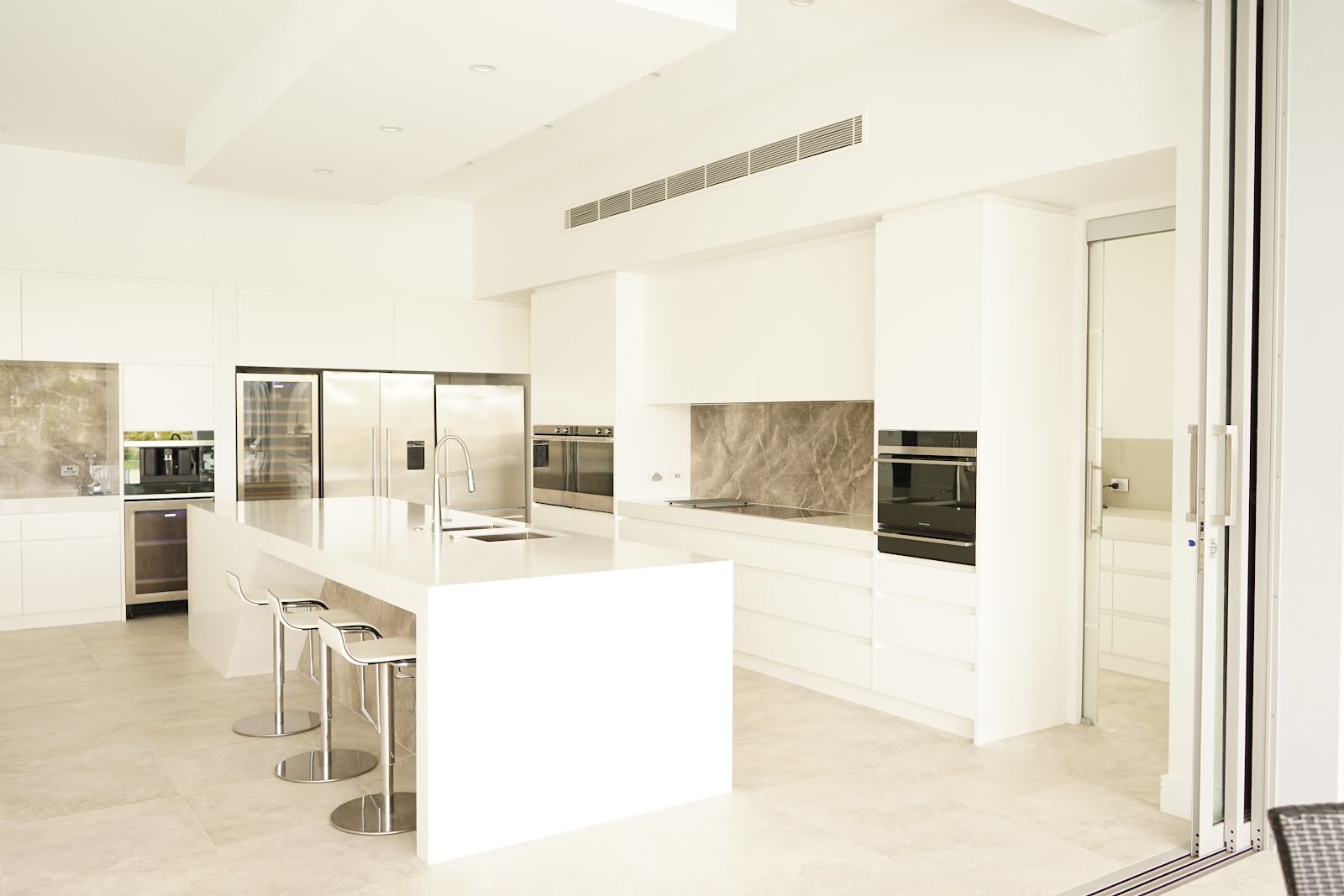 Bright white kitchen with a stone splashback, integrated ovens and bar stools
